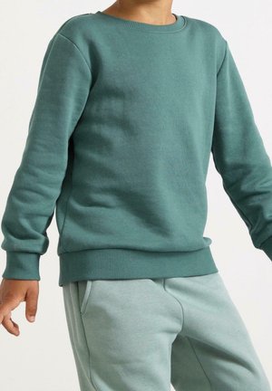 Niño vestido con sudadera de manga larga color verde azulado y pantalones deportivos azul claro grisáceos, con las manos relajadas a los lados sobre fondo blanco.