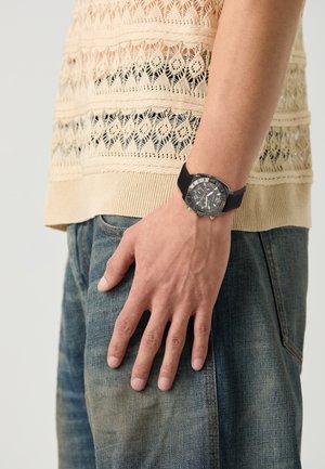 Svart og gull kronograf klokke med tre underdials og et teksturert belte, båret på håndleddet over denimjeans og en strikket beige topp.
