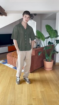 Kortærmet mørkegrøn skjorte med mønstret design, cremefarvede joggers og grå sneakers står på trægulv nær en plante.