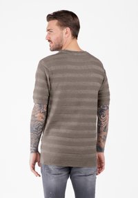 Uomo con braccia tatuate che indossa un maglione a maniche corte a righe taupe e jeans grigi strappati, rivolto a sinistra su uno sfondo semplice.