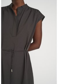 Ärmlös klänning i kolgrått med ståkrage, V-ringning och ett tunt midjeband på en person med mörk hy mot en enfärgad bakgrund.