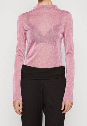 Kobieta w przezroczystym różowym topie z długim rękawem na pasujący biustonosz, w połączeniu z czarnymi spodniami z wysokim stanem, przedstawiona od szyi do bioder.