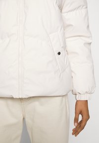 Zbliżenie na osobę w kremowej pikowanej kurtce z zamkiem błyskawicznym i kieszenią na zatrzask, w połączeniu z jasnym spodniami.