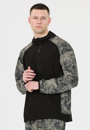 Jeune homme portant un sweat à capuche noir avec des manches grises à motifs abstraits et un pantalon assorti, debout sur un fond uni.
