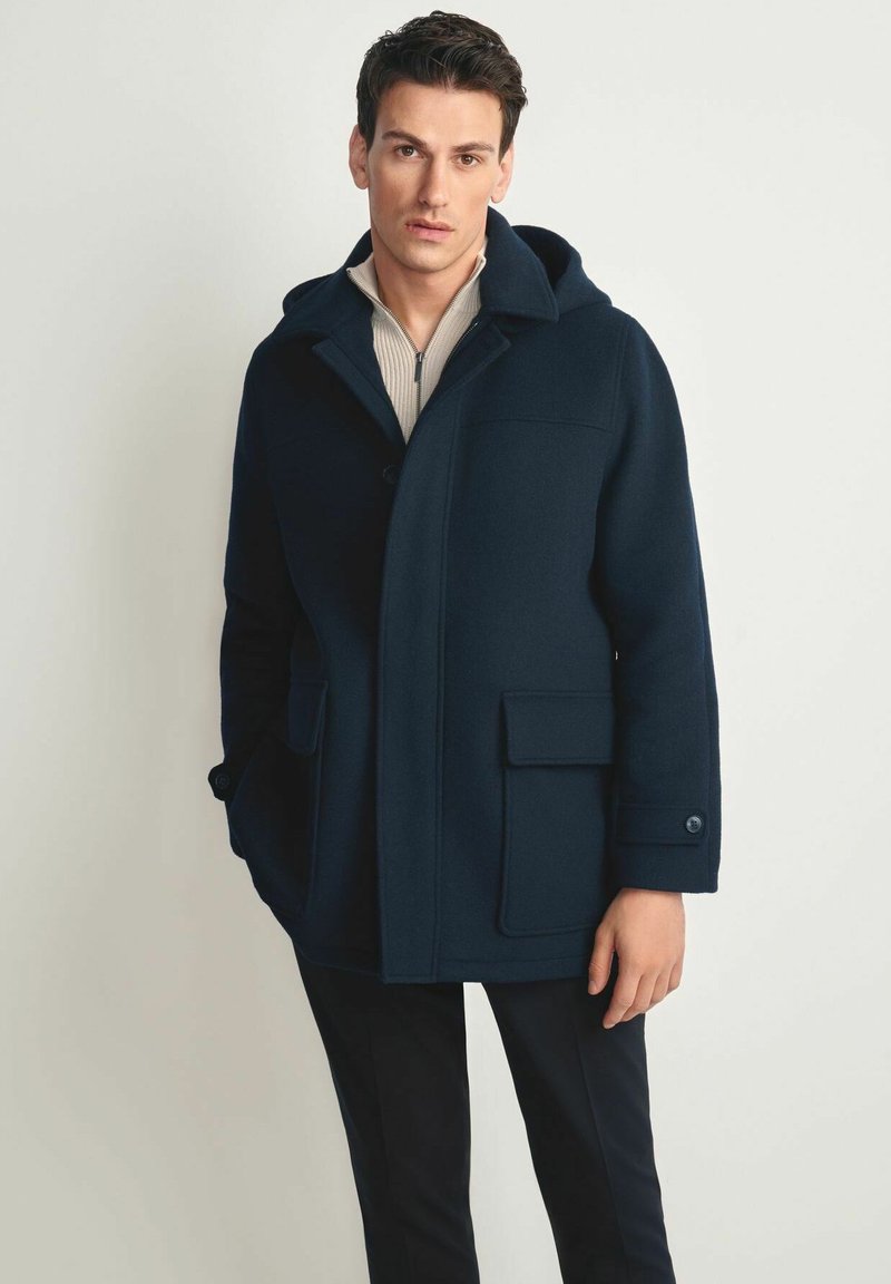 Granatowy wełniany płaszcz z kapturem, dużymi przednimi kieszeniami i zapięciem na guziki. Tkanina ma gładką fakturę, którą uzupełnia jasnobeżowa podszewka.