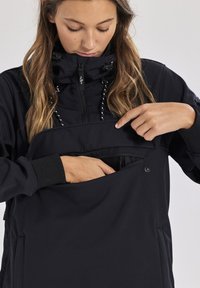 Svart softshell-jacka med hög krage, dragkedja fram och en stor framficka. Har snörningsdetaljer och ribbade ärmslut.