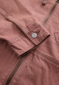 Gros plan sur la manche d'une veste en jean rose poussiéreux avec un bouton en métal et une fermeture éclair visible sur un tissu texturé.
