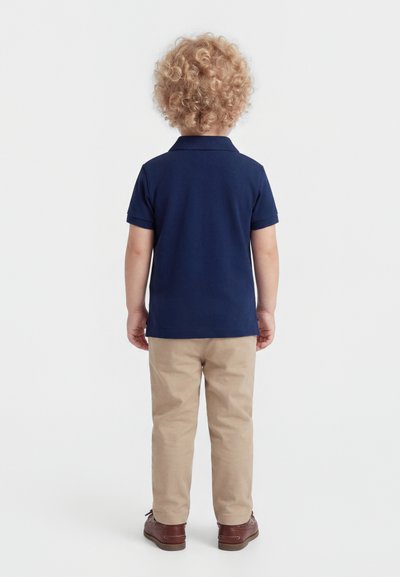 Malé dítě s kudrnatými blond vlasy stojící zády, oblečené v tmavě modrém polo tričku, béžových kalhotách a hnědých botách na bílém pozadí.