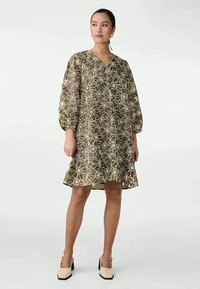 Vestido castanho com padrão floral, com decote em V, mangas bufantes e uma silhueta evasê. Feito de tecido leve, até ao joelho, combinado com sapatos de salto beiges.