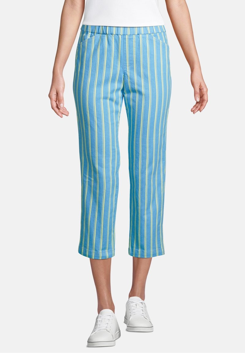 Femme portant un pantalon bleu court avec des rayures jaunes verticales et des baskets blanches, debout devant un fond uni.