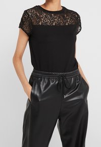Mujer vestida con una blusa de encaje negro con mangas cortas y pantalones de imitación de cuero negro con cordones, con las manos en los bolsillos.