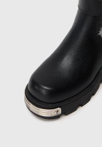 Bota de cuero negro con suela de tracción gruesa y acento metálico en la punta frontal sobre fondo blanco.
