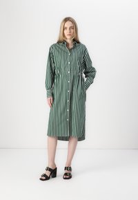 Rochie cămașă cu dungi verzi și albe, cu închidere buton, mâneci lungi, talie strânsă, până la genunchi, asortată cu sandale negre cu toc și catarame.
