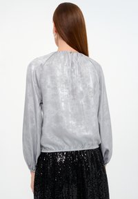 Silberne Bluse mit einer weichen, glänzenden Textur, gerafftem Ausschnitt und langen, puffigen Ärmeln. Elastischer Saum an der Taille, kombiniert mit einem glitzernden schwarzen Rock.