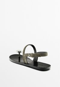 Sandal in suede grigia con suola nera piatta, dotata di una cinghia alla caviglia e di un accent metallico vicino all'alluce. Design minimalista con fibbia regolabile.