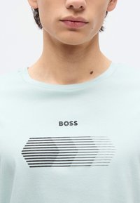 Νεαρό άτομο που φοράει ανοιχτό μπλε πουκάμισο με τη λέξη "BOSS" και μια μαύρη γεωμετρική γραμμή σχεδιασμένη στο κέντρο κάτω από τη γραμμή του λαιμού.