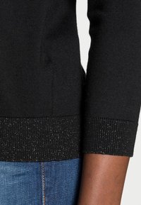 Schwarzer Pullover mit strukturiertem Finish und tailliertem Schnitt. Die Bündchen verfügen über ein glitzerndes, geripptes Detail. Kombiniert mit blauen Denim-Jeans.