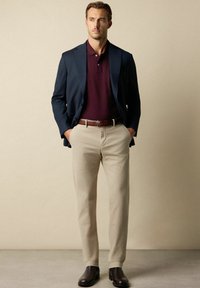 Granatowa marynarka na granatowym polu, sparowana z jasnobeżowymi spodniami i ciemnobrązowymi butami. Gładkie materiały o dopasowanym kroju.