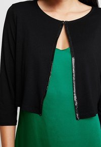 Mustat lyhyet neuleet, joissa on kolme neljännestä hihoista ja etulukko, sekä kiiltävä helmirivistö keskellä, päällä vihreä mekko.