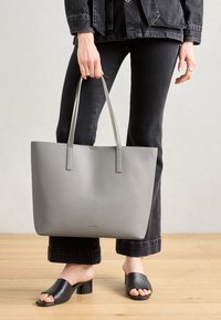 Siva torba s dva duga ručka, glatke teksture, minimalističkog dizajna, zlatni logo sprijeda, u kombinaciji s crnim trapericama i sandalama.