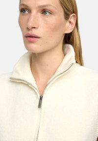 Kremowy sweter zapinany na zamek o teksturowanej, miękkiej tkaninie. Wysoki kołnierz i metalowy zamek podkreślają design. Minimalistyczny i przytulny wygląd.