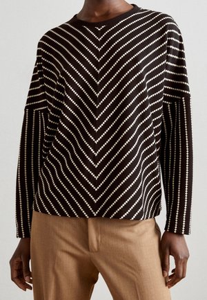 Pull rayé noir et blanc avec motif en chevron, associé à un pantalon marron clair, porté par une personne debout devant un fond uni.