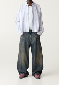 Jasnoniebieska, strukturalna koszula z guzikiem na biały T-shirt, w połączeniu z szerokimi, wyblakłymi niebieskimi dżinsami i różowymi trampkami. Widoczny detal ze srebrnym łańcuszkiem.