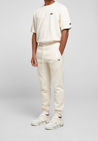 Camiseta de algodón crema con un pequeño logotipo en el pecho izquierdo, combinada con pantalones de chándal crema a juego que cuentan con bolsillos laterales y una cinturilla ajustable.