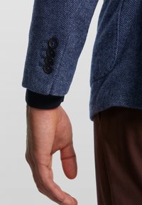 Mörkblå herringbone-jacka med en ärm prydd av fyra svarta knappar och en ribbad marinblå manschett; hand som vilar nedanför bruna byxor.