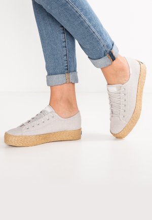 Person iført lysegrå lærredssneakers med vævede rebsåler og opkrogede blå jeans mod en ensfarvet hvid baggrund.