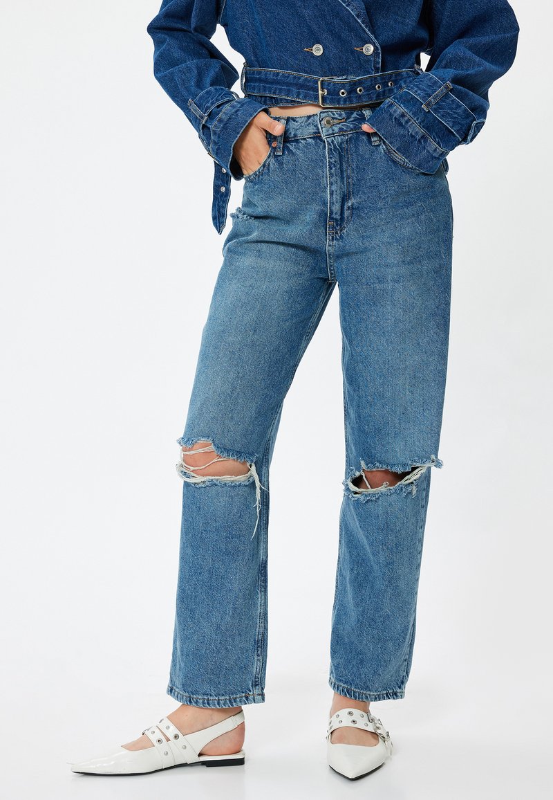 Jeans de mezclilla azul claro rasgados con un corte holgado, con bordes deshilachados en las rodillas y una cintura alta, combinados con zapatos planos blancos de punta.