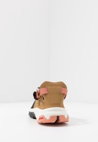 Scarpa da trail con tomaia sintetica marrone, pannelli laterali neri e dettagli rosa. Presenta un colletto imbottito e una suola bianca e testurizzata.