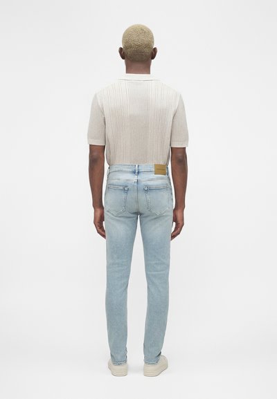 Άνδρας με κοντά ξανθά μαλλιά, που φοράει μπεζ κοντομάνικο ριμπ πόλο πουκάμισο, ανοιχτό μπλε τζιν και λευκά αθλητικά παπούτσια, στέκεται γυρισμένος από την πλάτη.