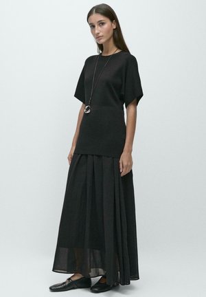 Femme aux longs cheveux foncés portant un haut noir ample à manches courtes, une longue jupe noire plissée, des ballerines noires et un long collier avec un pendentif circulaire.