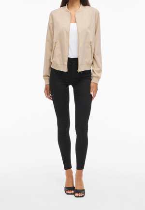 Kvinde iført beige bomberjakke, hvid top, sorte skinny bukser og sorte åbne stiletter står på en hvid baggrund.