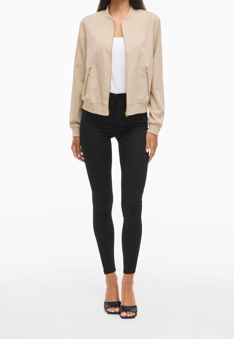 Femme portant une veste bomber beige, un haut blanc, un pantalon noir moulant et des talons ouverts noirs, debout sur fond blanc.