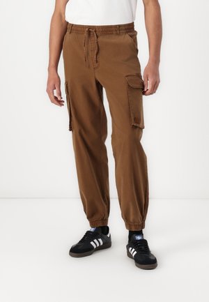 Hombre con pantalones cargo marrones con puños elásticos y cintura con cordón, zapatillas negras Adidas con rayas blancas, de pie frente a un fondo liso.