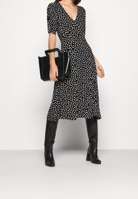 Černé midi šaty s puntíky, krátkými nafouknutými rukávy, véčkovým výstřihem a splývavou sukní, doplněné lesklou černou obdélníkovou kabelkou a vysokými botami po kolena.