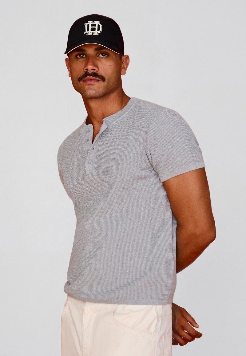 Homme avec moustache portant une casquette noire, une chemise grise à manches courtes boutonnée et un pantalon clair, debout devant un fond uni.