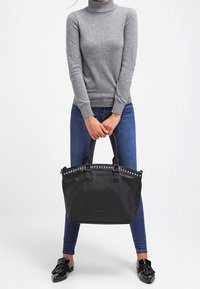 Person i grå polotröja och blå jeans som håller en stor svart handväska med nitar och svarta tofflor med fransar.