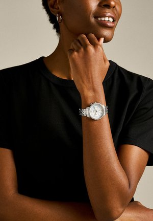 Person iført sort skjorte med kort hår smiler, hviler hagen på hånden og bærer et sølvfarvet metal-link chronograph armbåndsur med rhinsten.