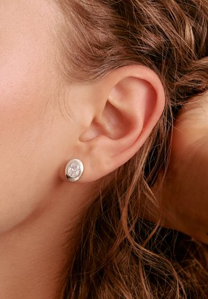 Nahaufnahme eines Ohrs mit welligem braunem Haar, das einen kleinen, runden silbernen Ohrring mit einem klaren Edelstein trägt.