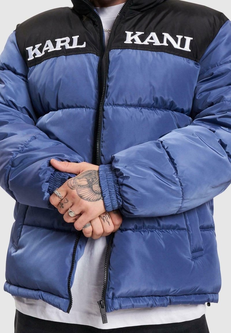 Puffjacka i blått med en svart överdel. Har kviltad textur, dragkedja fram och broderad "KARL KANI"-logga på bröstet.