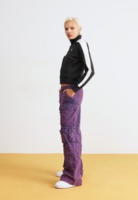 Black zip-up jacket with white stripes, paired with purple cargo pants featuring multiple pockets and a relaxed fit, complemented by white sneakers.