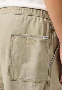 Beżowe szorty z naszywaną kieszenią i niebieskimi detalami, z metką marki Wrangler na kieszeni. Miękka, teksturowana tkanina.