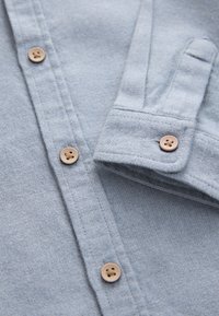 Camicia in tessuto azzurro chiaro con trama, colletto con bottoni e bottoni marroni con quattro fori. Vista ravvicinata che mette in evidenza i dettagli delle cuciture.