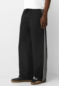 Mannelijke model in zwarte wijde broek met grijze zijpanelen en witte bies, gecombineerd met zwarte sneakers met rubberen zolen en witte veters.