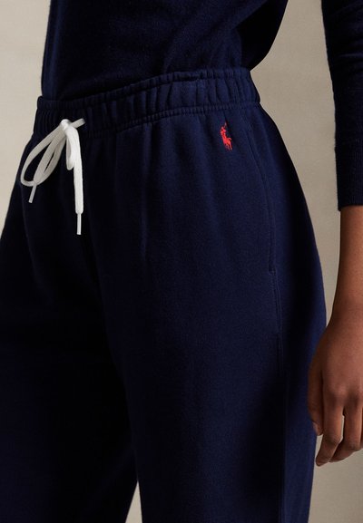 Marineblauwe joggingbroek met wit trekkoord, rood geborduurd logo op de heup, gedragen met bijpassende marineblauwe trui, staand tegen een neutrale achtergrond.