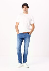 Camiseta de algodón blanca, jeans de mezclilla azules, cinturón marrón y zapatillas blancas. El modelo está de pie con las manos en los bolsillos contra un fondo liso.