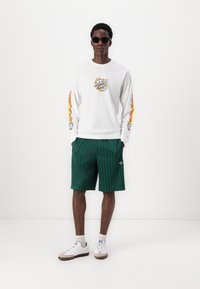 Long-sleeve white shirt with flame graphic and green striped shorts. Model wears black sunglasses and white sneakers with brown accents.